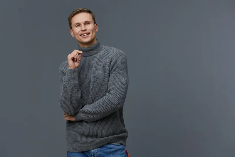 smiling young caucasian man wearing gray knitted turtleneck sweater and blue jeans look camera hold hand near face isolated on gray background casual style concept digital nomad lifestyle