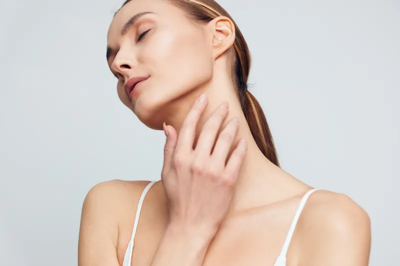 A young woman with closed eyes gently touches her neck, showcasing smooth skin and a calm expression against a plain light background. Concept for beauty and skincare.