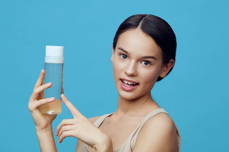 Young woman holds skincare product, showcasing healthy skin and confidence against a vibrant blue background, emphasizing beauty and self care