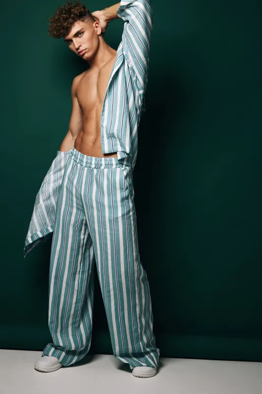 Young man posing in striped loose pants and open shirt with bare chest against dark background. Fashion model with curly hair and casual style in studio photo shoot.
