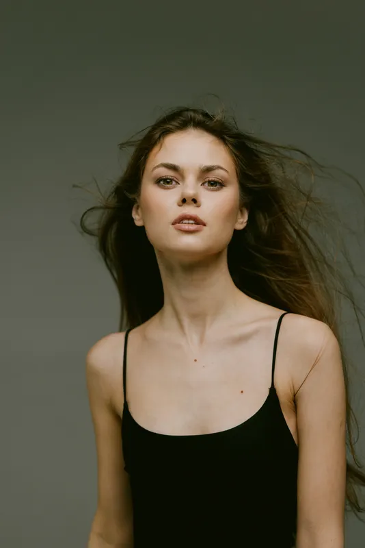 Young woman with long hair wearing black top looking confident and natural with tousled hair against gray background