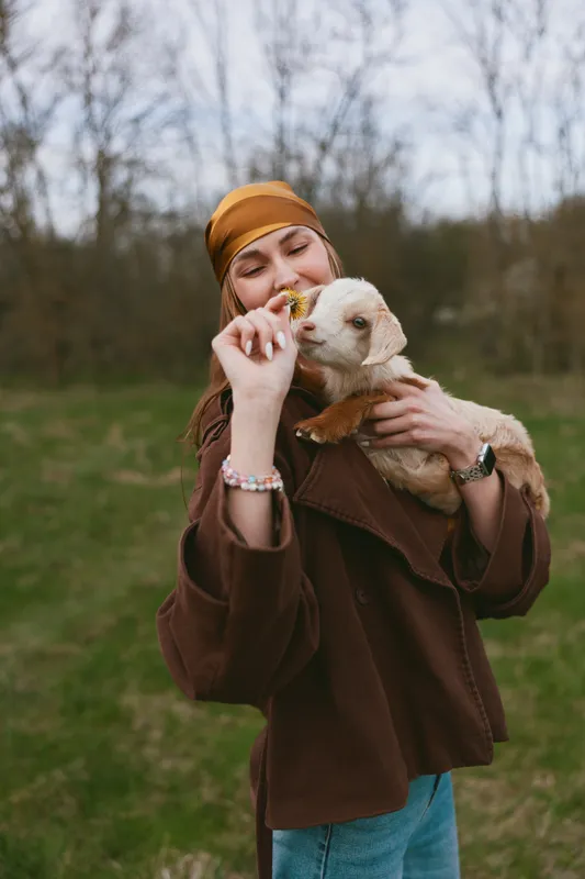 Smiling woman in a brown coat and mustard hat feeds a curious goat kid outdoors. Natural light, park setting, lifestyle moment highlighting companionship between human and animal.