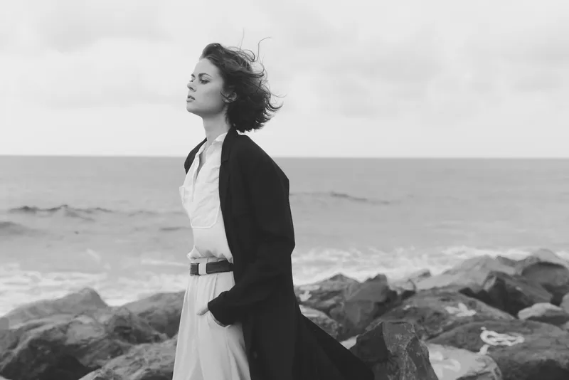 A windswept woman stands on jagged rocks by the sea, wearing a long coat over a light dress, expressing resolve and calm against a gray horizon.