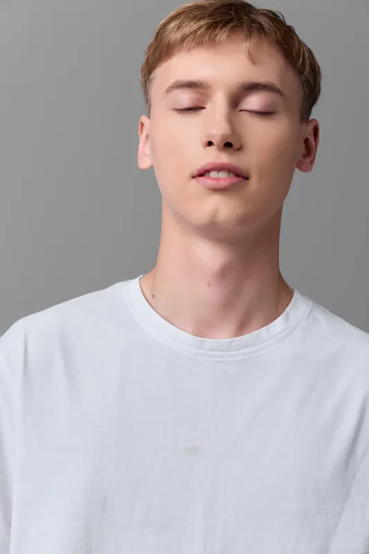 Young man with closed eyes wearing a white shirt conveys calm, relaxation and serenity in a minimal studio setting, emphasizing inner balance, mindfulness, and comfortable stillness
