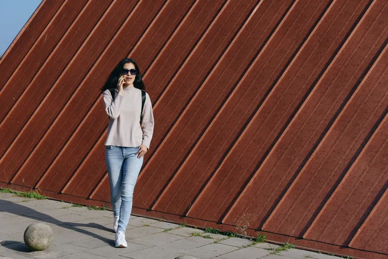 Woman in sunglasses, light sweater and jeans walks beside a diagonal brown wall in an outdoor urban setting, showcasing casual street style and relaxed pose.
