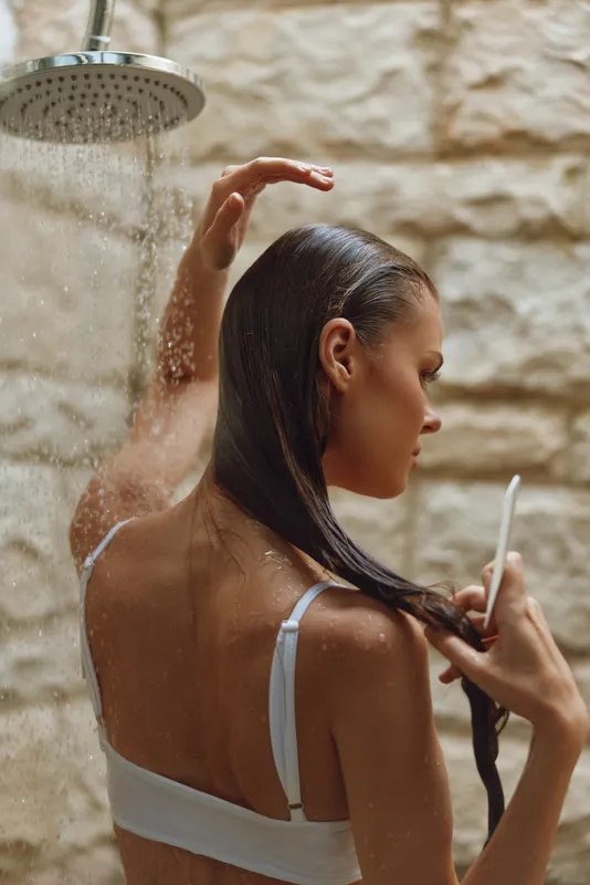 Woman showering outdoors under rain shower, wet long hair, natural stone wall, relaxed morning routine, casual white underwear, soft natural light.