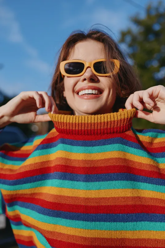 Woman smiling in a colorful striped sweater and yellow sunglasses outdoors, young adult joyful in a casual outfit, pulling a turtleneck in sunshine for a playful lifestyle fashion portrait.
