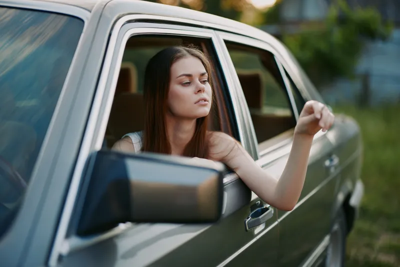A young woman with long hair enjoying the breeze while sitting in a vintage car, showcasing a relaxed mood in a natural outdoor setting.