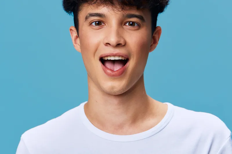 smiling man with curly hair wearing white t-shirt on blue background, expressing surprise and joy, casual young adult portrait with vibrant colors