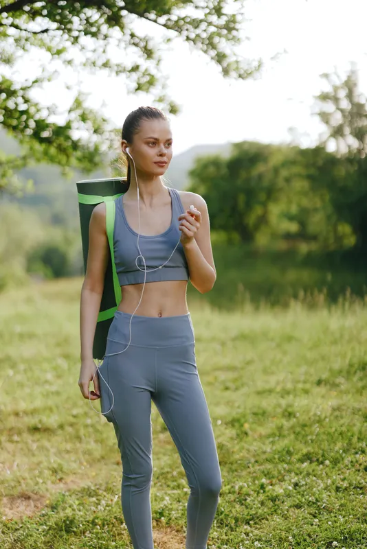 Young woman in sportswear enjoying a moment outdoors with a yoga mat, symbolizing health and wellness in nature, featuring fresh green tones