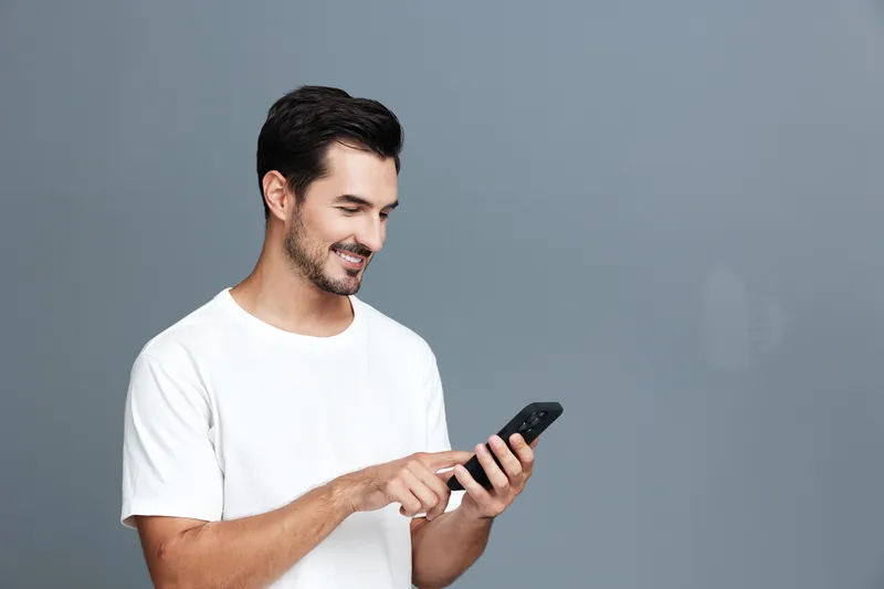Smiling young man wearing white t-shirt looking at mobile phone and enjoying his time, isolated on a soft grey background studio portrait. People lifestyle concept.