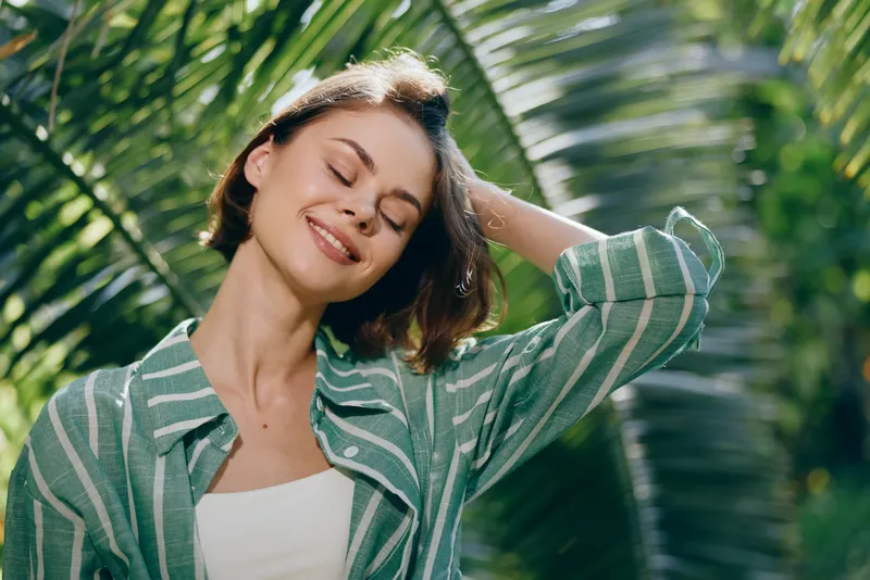 Joyful woman outdoors, sunlit smile, wearing a green striped jacket, relaxed pose amid lush tropical foliage and warm natural light.