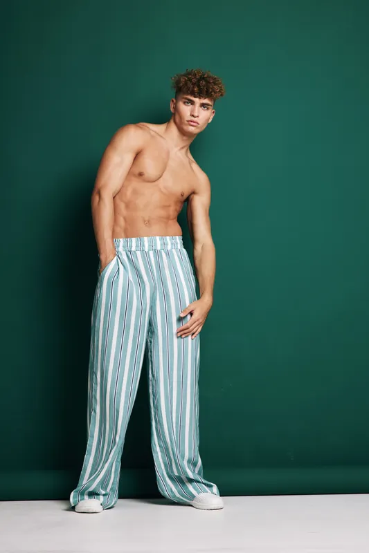 Young man with curly hair posing shirtless in striped wide pants and white sneakers against green background. Fashion model standing with hands in pockets and serious expression.
