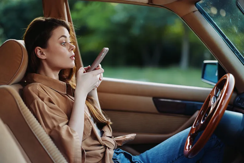 Young woman in a beige shirt using her smartphone while relaxing in a car interior, with a green background, representing modern lifestyle and technology.
