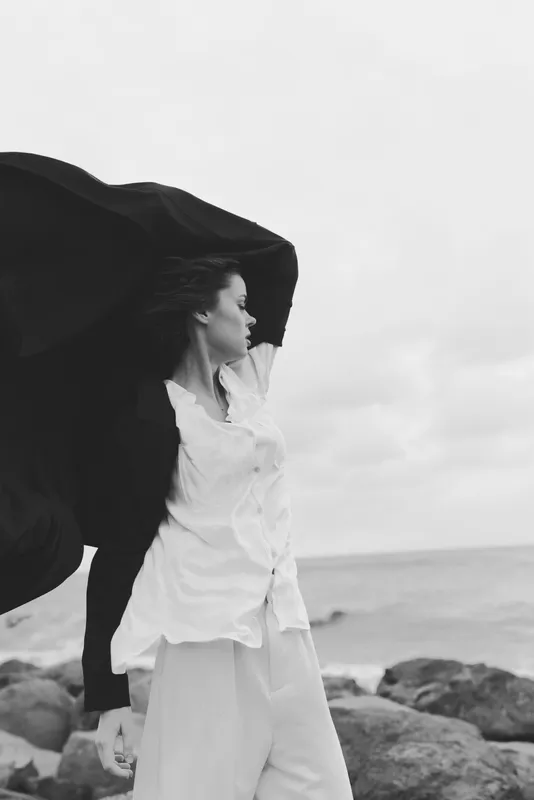 Wind sweeps a cape as a woman in white stands on a rocky coast by the sea, black and white photography capturing motion, freedom and coastal fashion in a dramatic moment