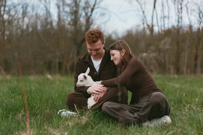 A couple outdoors in a green field cuddle a white goat kid, capturing tender bonding moments. Natural light, casual clothing, and rural scenery convey everyday affection, playfulness, and connection