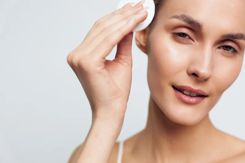 Close-up of a young woman cleansing her face with a cotton pad on a white background. Concept of skincare, beauty routine, self-care, clear skin, and healthy lifestyle.