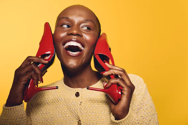 Smiling black woman with dark skin holds red high heels near her face, wearing beige sweater. Bright yellow background highlights joyful mood and fashion concept with stylish accessories.