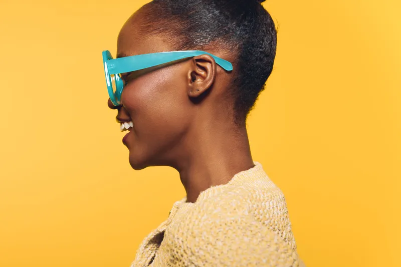 Profile of a smiling dark skinned African woman wearing large blue glasses and a beige sweater against a bright yellow background. Concept of fashion, happiness, and vibrant lifestyle.