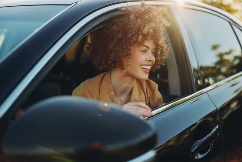 Woman leans from a car window, smiling, enjoying a bright moment on a sunny drive, lifestyle scene with retro film color and candid joy, casual fashion and travel mood