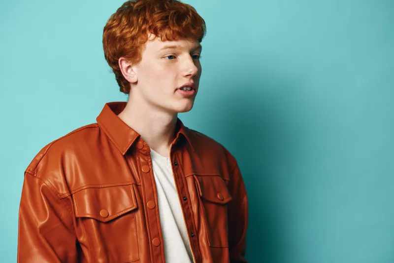 Young man with red hair wearing a brown leather jacket and white shirt standing against blue background looking sideways with neutral expression and casual style.