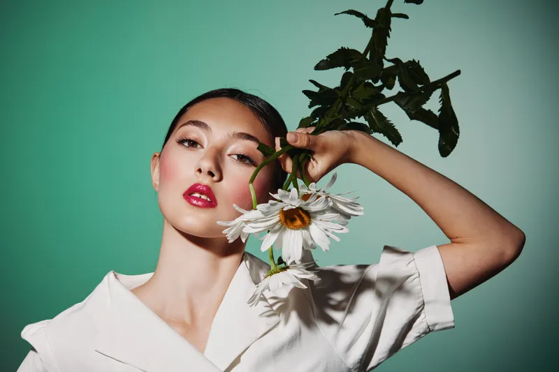 Fashion model with glossy red lips holds drooping white flowers against green background. Creative beauty shot with soft lighting and iconic stylized pose and expression.