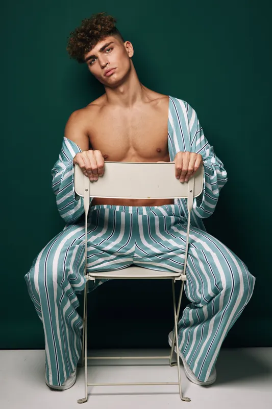 Young man with curly hair wearing striped pajamas sitting backwards on white chair against dark green background in studio portrait fashion concept.