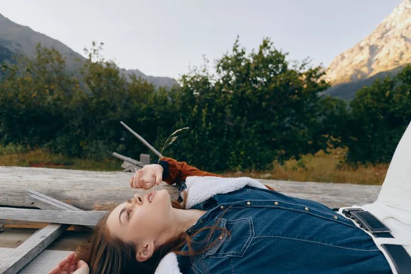 Woman relaxation outdoors in mountains, denim jacket portrait of a young model lying on wooden bench by forest, nature scenery conveying peaceful resting, leisure and calm.