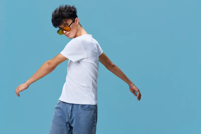 young man wearing sunglasses in casual white t-shirt and jeans posing confidently against blue background with curly hair, modern style, summer vibes