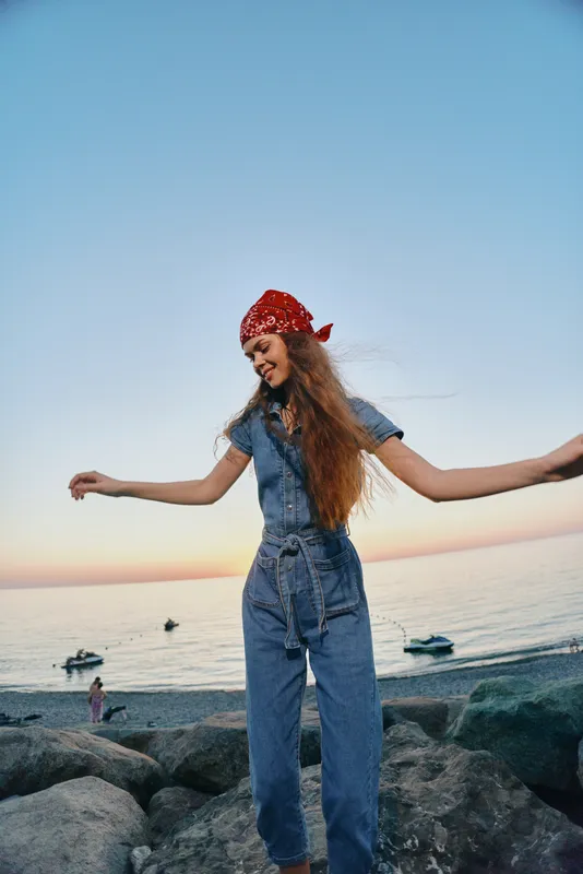 Lifestyle woman in modern boho-western denim street style smiling outdoors by the seaside during a film color sunset, casual and free-spirited mood.