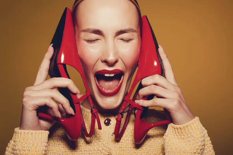 Excited woman with closed eyes holds red high heels near face, wearing yellow sweater. Concept of fashion, style, happiness, and vibrant emotions in studio with warm background.