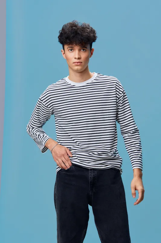 Young man wearing a striped long sleeve shirt and black pants stands against a plain blue background with a serious expression, casual fashion style.