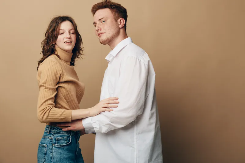 Young couple embraces gently against beige background. Woman in brown sweater and jeans closes eyes while man in white shirt looks at camera. Concept of affection and intimacy.