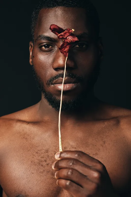 African man with dark skin holding dried red flower in front of face, creative conceptual portrait with visual style and artistic expression on black background.