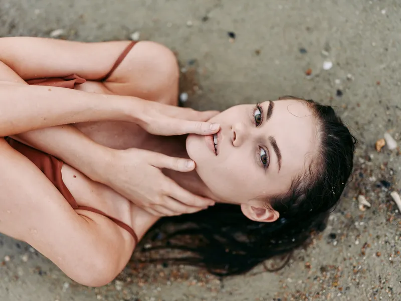 Portrait of a woman lying on a sandy beach with wet hair spread around, soft gaze, and hands framing her face, conveying calm, vulnerability, and natural beauty.