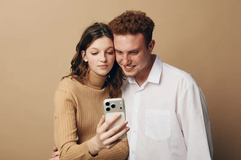 Young couple with curly hair takes selfie together using smartphone on beige background. They smile and enjoy moment, showing affection and modern lifestyle with technology.