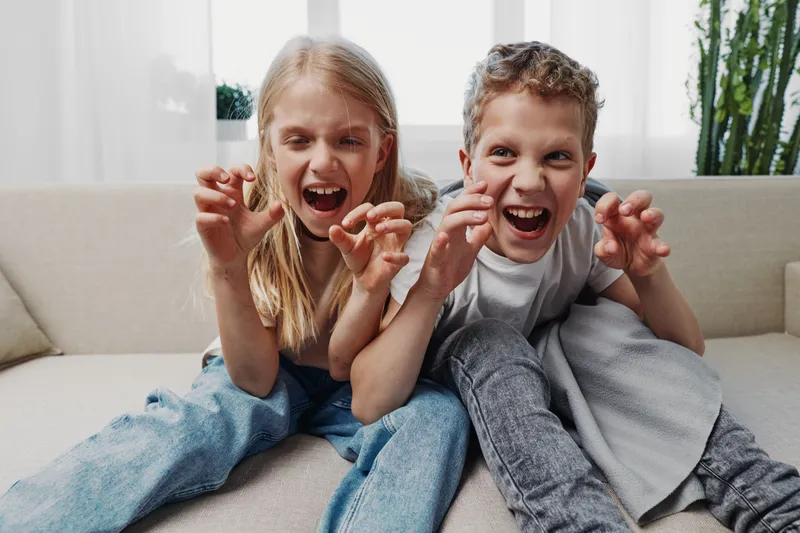 Two playful children sitting on a sofa make funny claw gestures and faces while enjoying a bright cozy room. Concept for childhood fun and sibling bonding.