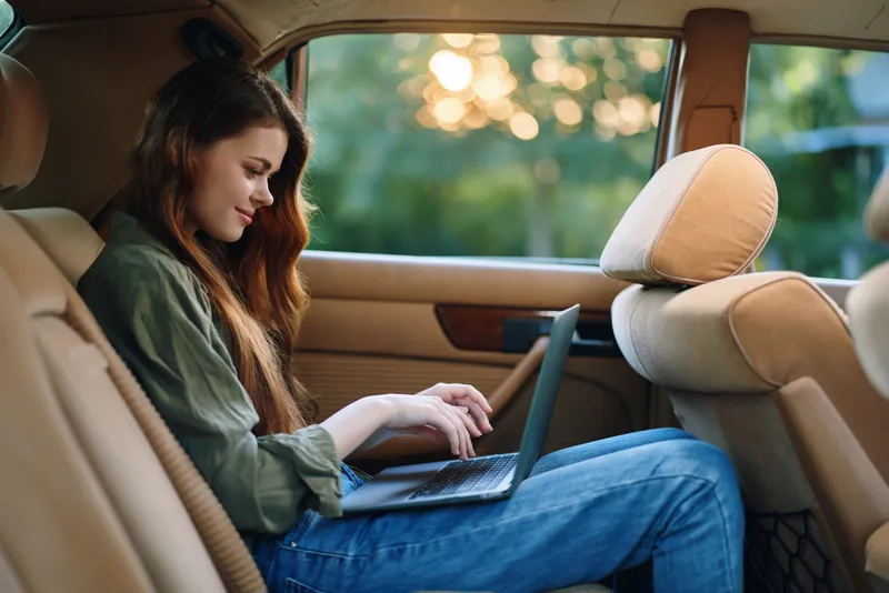 Young woman using a laptop inside a car, enjoying the outdoor view, showcasing a relaxed and productive lifestyle with green nature background.