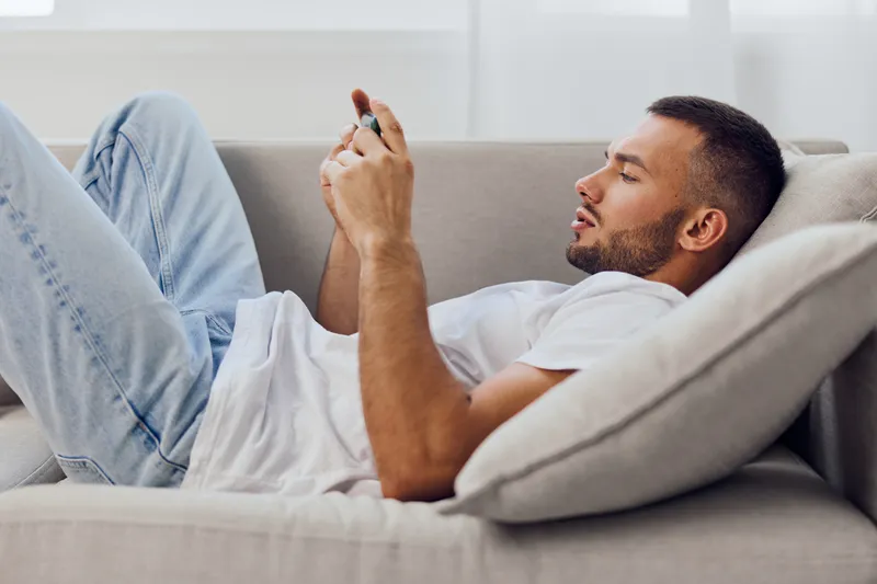 Casual man relaxing on a sofa, focused on his smartphone, the bright living room exuding comfort and modern style. A perfect blend of leisure and technology.