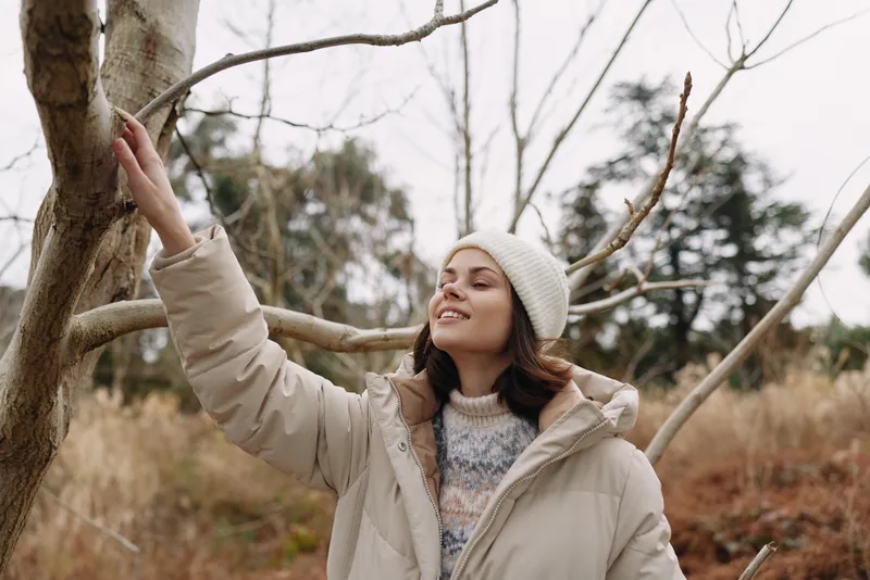 Woman outdoors in winterwear touches a bare tree branch, smiling amid a natural landscape with soft light, wearing a cozy jacket and beanie to convey calm, warmth, and seasonal serenity