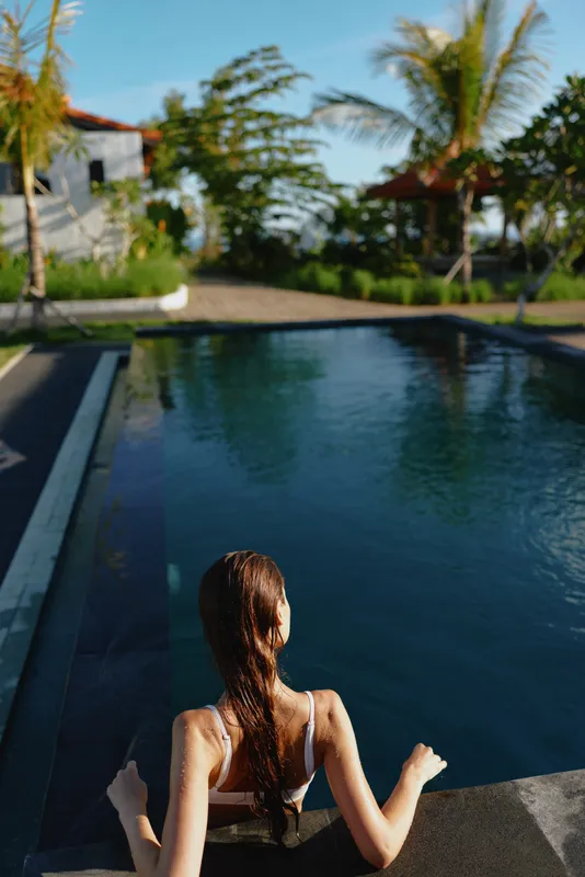 A woman in a swimsuit approaches a bright resort pool, capturing a carefree lifestyle moment by the water with tropical palms and modern cabanas in the background.