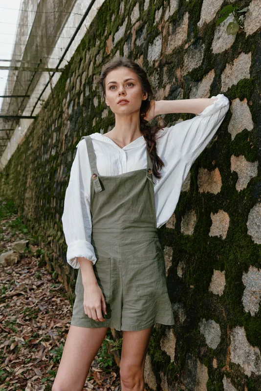 Young woman in casual summer outfit, featuring a white shirt and olive green overalls, posing against a rustic stone wall in a natural setting, conveying confidence and tranquility
