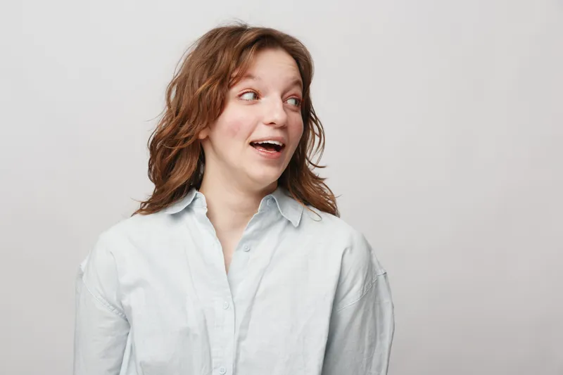 Young woman with curly hair wearing white shirt looks surprised and happy against light background. Concept of positive emotions, casual style, and natural beauty in studio portrait.
