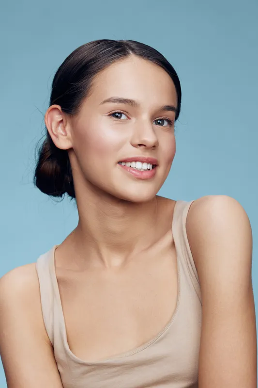 A smiling young woman in a simple beige tank top against a light blue background, showcasing natural beauty and a fresh look ideal for health and beauty concepts