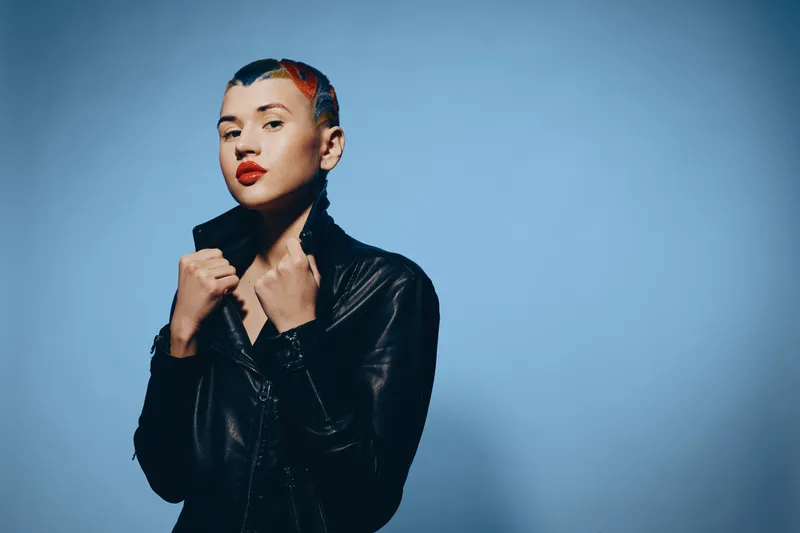 Bold woman with colorful short hair wearing a black leather jacket confidently poses against a smooth blue background, showing style and attitude in modern fashion.