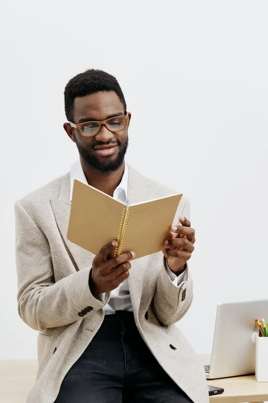 Professional young African-American man dressed in smart casual, reading a notebook with a slight smile in an office environment, wearing glasses, calm and focused expression, white background, modern