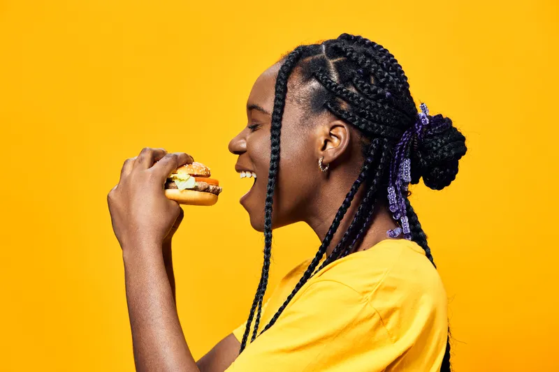 Cheerful young Black woman with braided hair enjoying a delicious hamburger against a vibrant yellow background, showcasing a love for healthy eating and diversity