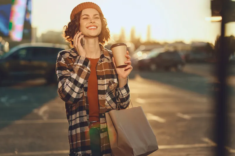 woman coffee phone smile shopping urban golden - Young woman in plaid coat enjoys coffee and talks on phone with a bright smile, shopping bag in hand at golden hour glow, authenticity and candid