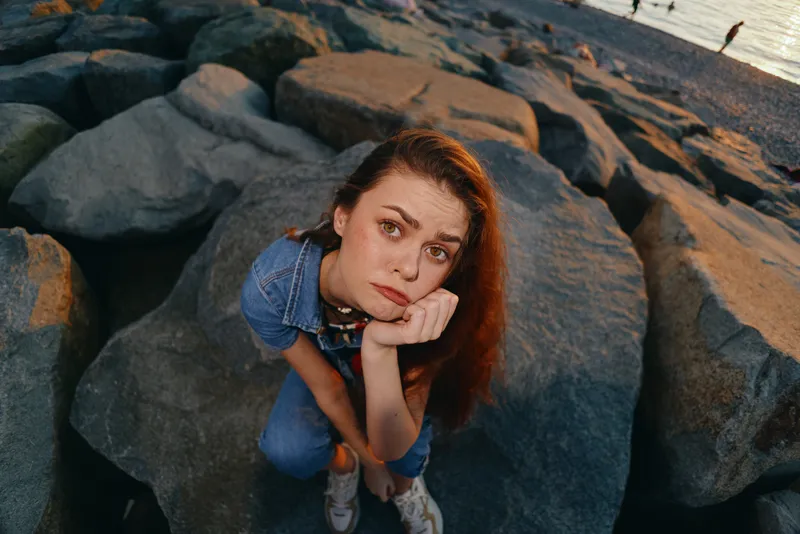 lifestyle woman in modern boho-western denim street style with thoughtful expression sitting on rocks, film color aesthetic capturing natural light mood and casual vibe