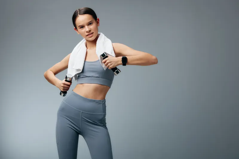 Woman in gray athletic wear with a towel on her shoulders performs a dynamic workout with dumbbells in a studio, showcasing fitness, strength, and focused training.